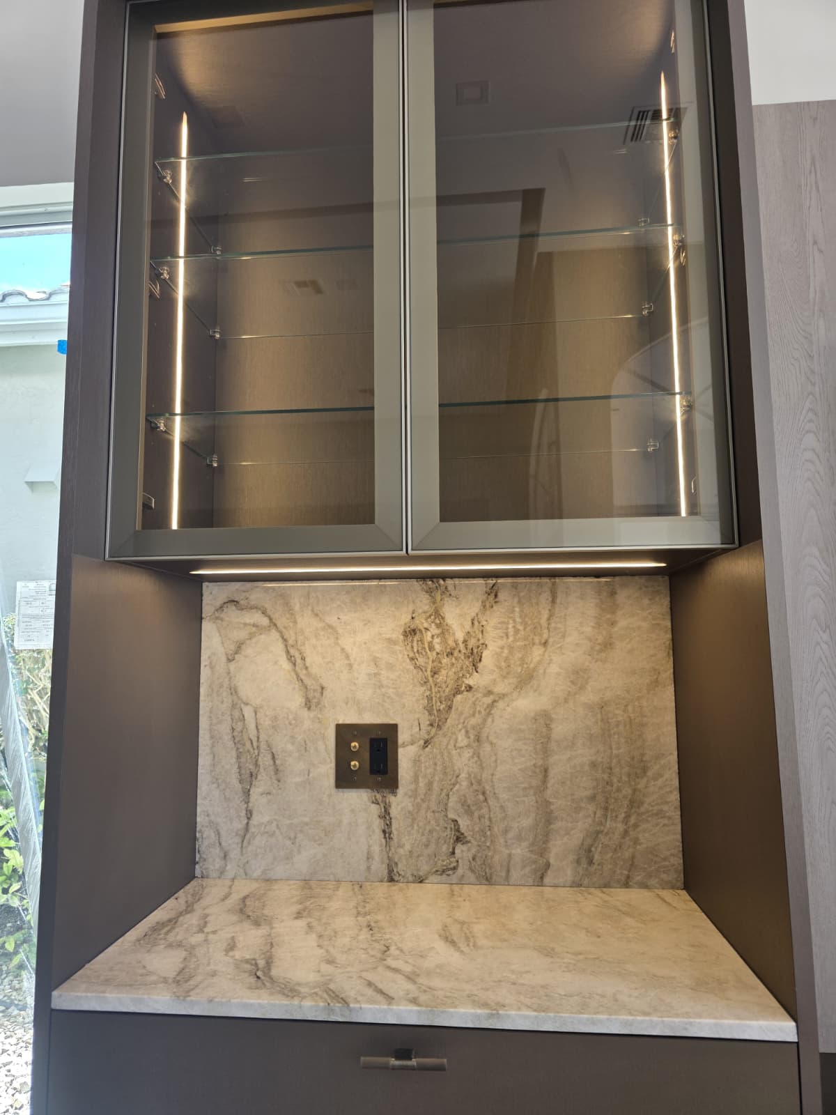 Glass-front cabinet with vertical LED strips, under-cabinet lighting, and marble backsplash with outlet.