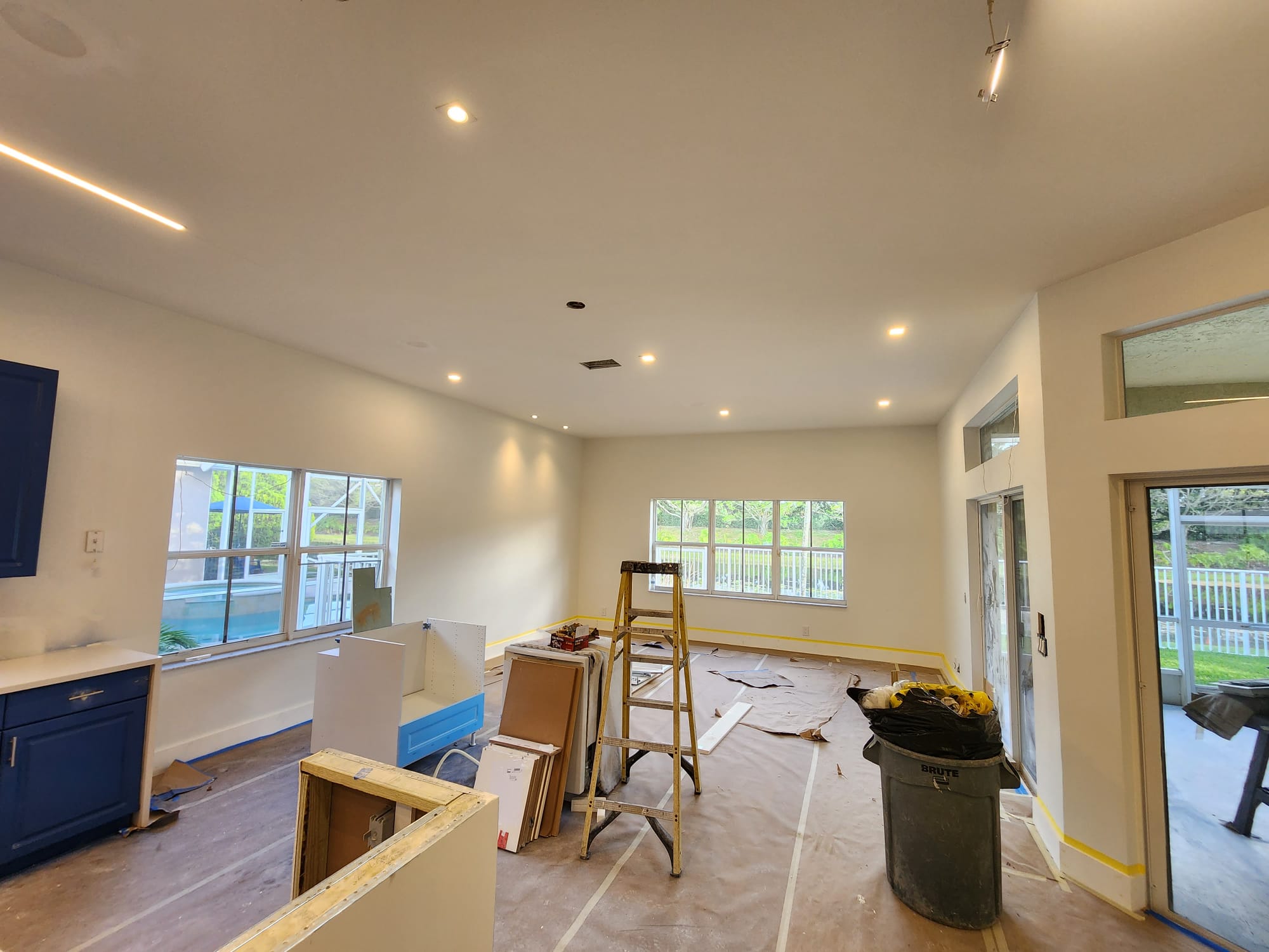 Room under renovation with warm recessed ceiling lights, linear LED strip, ladder, and kitchen cabinets.