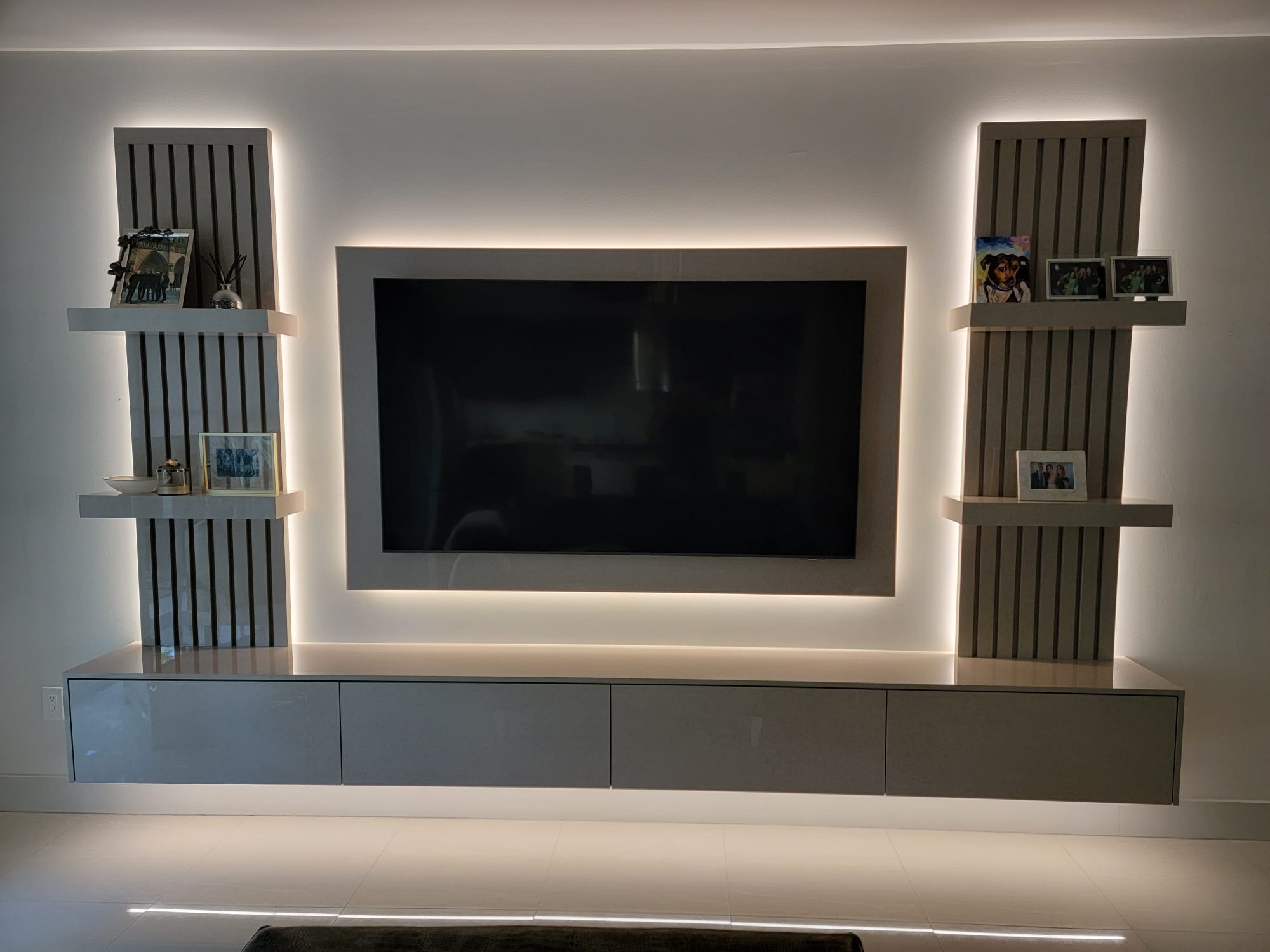 Symmetrical media wall with backlit TV frame, wood-slat side panels, floating shelves, and under-cabinet glow.