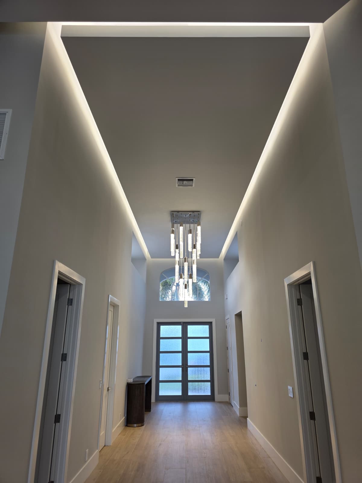 Tall hallway with tray-ceiling cove LEDs, cascading glass pendant, and frosted double doors.