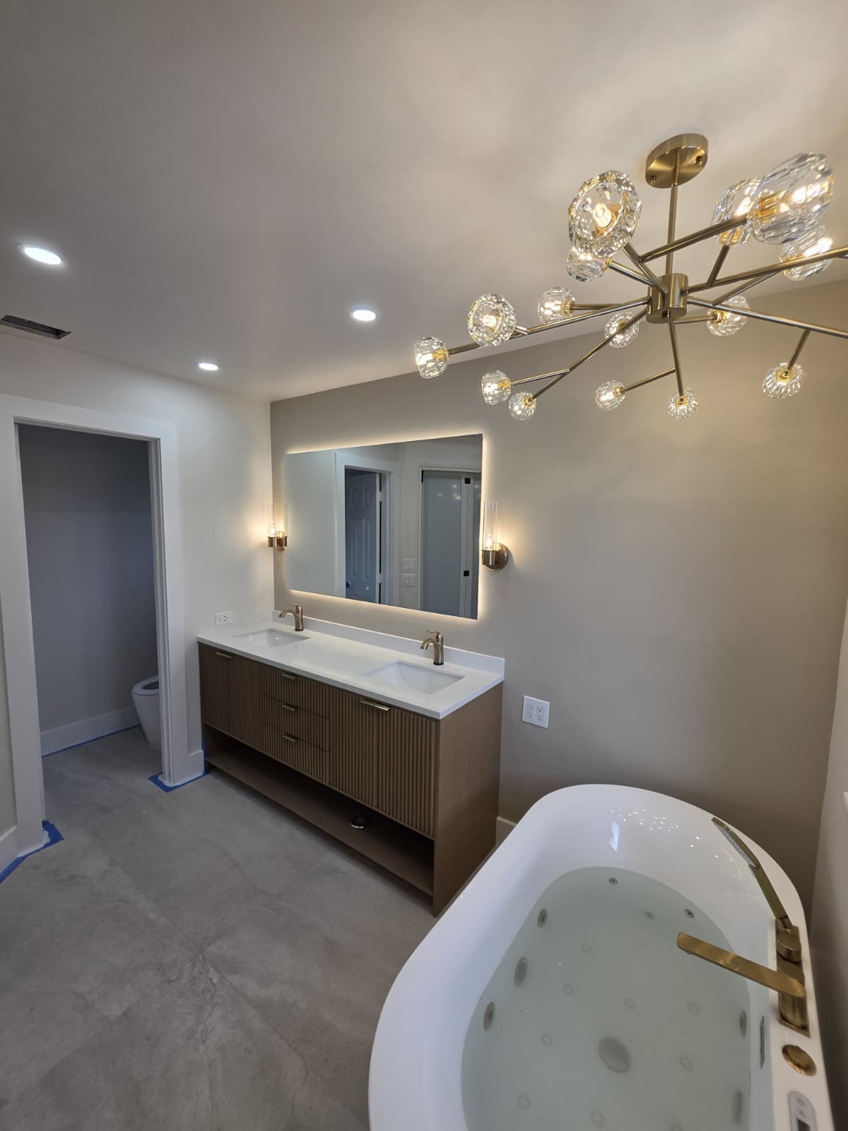 Bathroom with gold chandelier, LED-backlit rectangular mirror, sconces, and a light-wood double vanity.