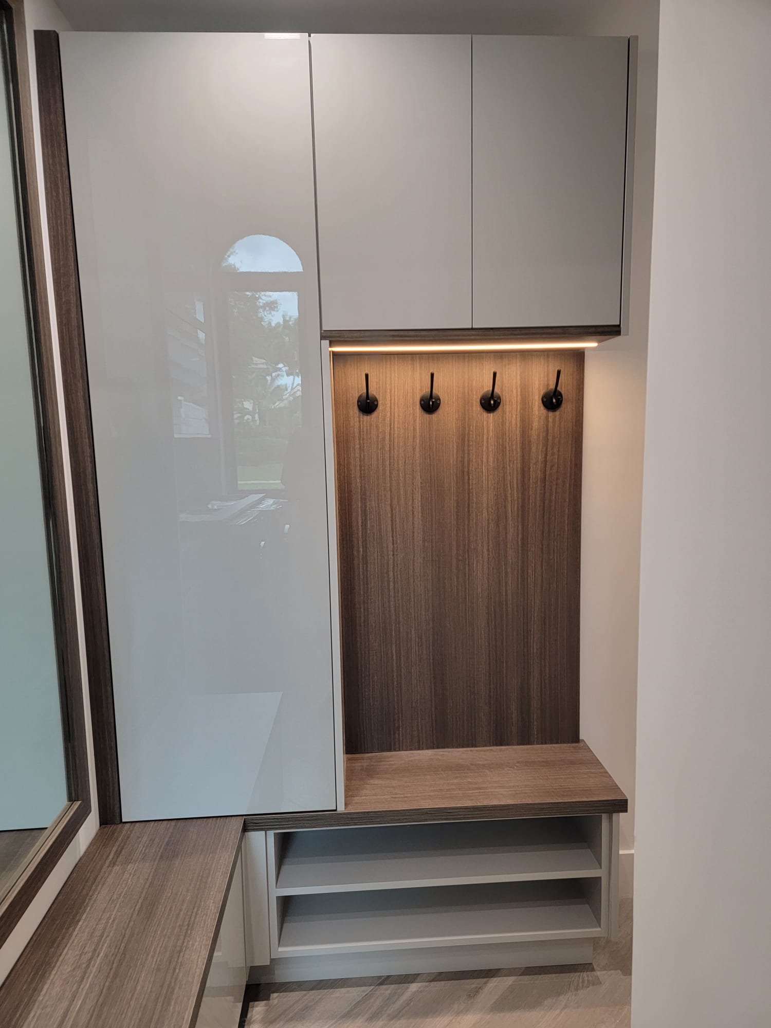 Modern entryway cabinet with glossy gray doors, wood alcove, black hooks, and under-cabinet LED accent lighting.